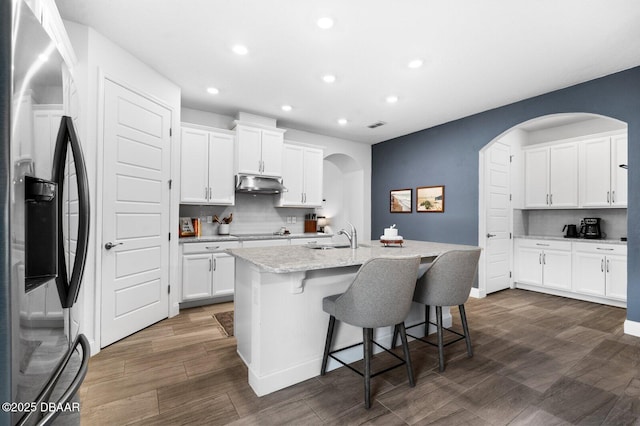 kitchen with black electric stovetop, under cabinet range hood, white cabinets, stainless steel fridge with ice dispenser, and a center island with sink