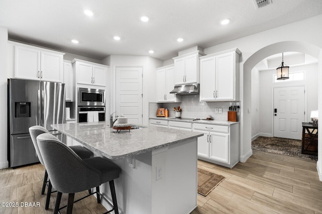 kitchen featuring arched walkways, a breakfast bar area, stainless steel appliances, tasteful backsplash, and under cabinet range hood