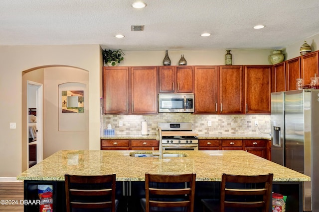 kitchen with open shelves, tasteful backsplash, appliances with stainless steel finishes, an island with sink, and light stone countertops