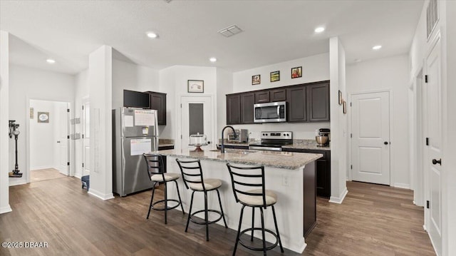 kitchen featuring visible vents, an island with sink, appliances with stainless steel finishes, wood finished floors, and a sink