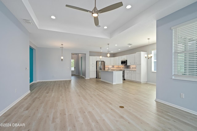 unfurnished living room featuring a tray ceiling, visible vents, light wood-style flooring, baseboards, and ceiling fan with notable chandelier