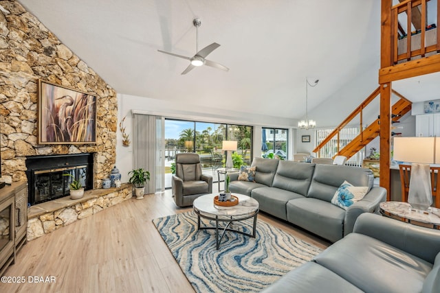 living room featuring high vaulted ceiling, ceiling fan with notable chandelier, a stone fireplace, and light hardwood / wood-style flooring