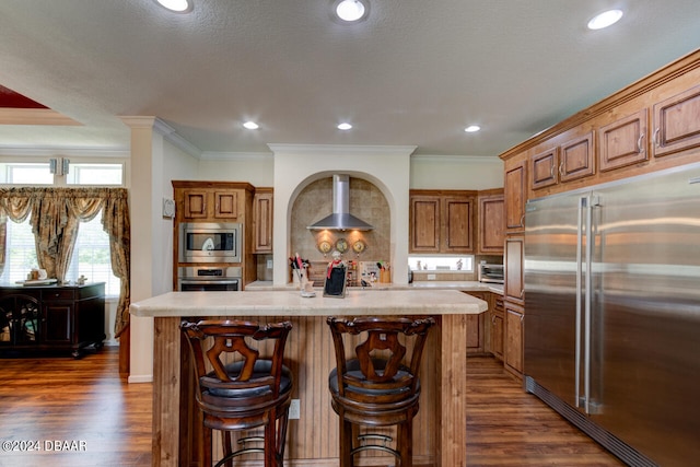 kitchen featuring a center island with sink, crown molding, wall chimney range hood, built in appliances, and dark wood-type flooring