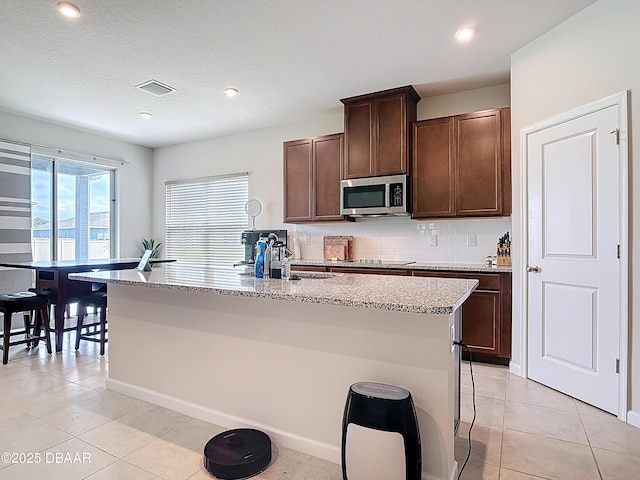 kitchen with stainless steel microwave, visible vents, an island with sink, decorative backsplash, and light tile patterned flooring