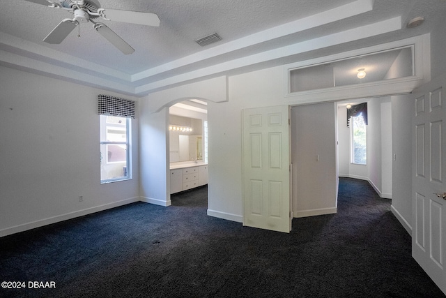 unfurnished bedroom featuring ensuite bath, ceiling fan, a raised ceiling, and dark carpet