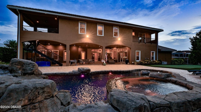 back house at dusk featuring a patio area, a balcony, and a pool with hot tub