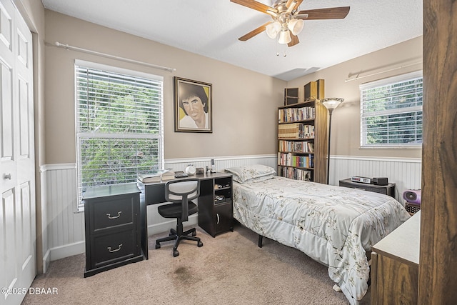 carpeted bedroom with a wainscoted wall, multiple windows, an office area, a textured ceiling, and ceiling fan