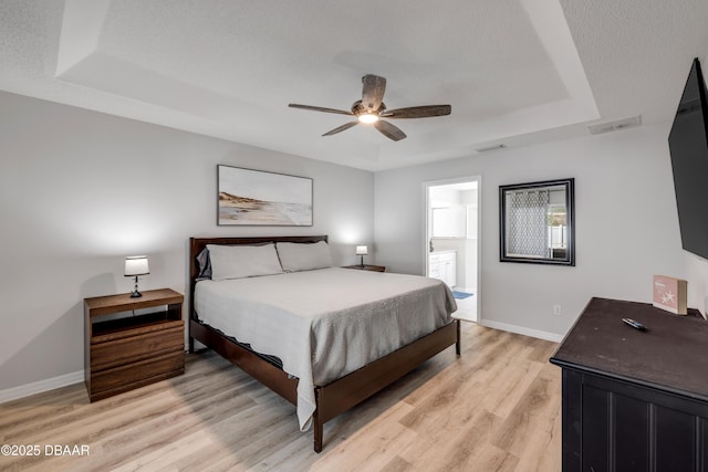 bedroom featuring a ceiling fan, baseboards, ensuite bath, a tray ceiling, and light wood-style floors