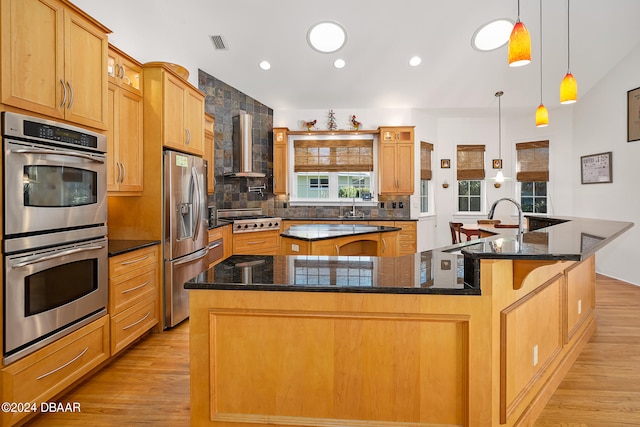 kitchen with stainless steel appliances, light hardwood / wood-style floors, wall chimney exhaust hood, hanging light fixtures, and a large island with sink