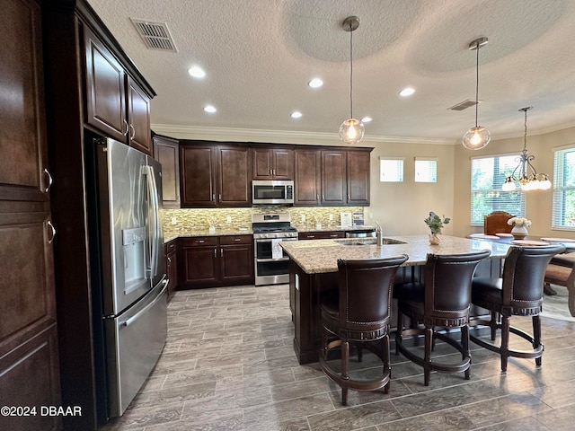 kitchen with appliances with stainless steel finishes, light stone counters, sink, pendant lighting, and an island with sink