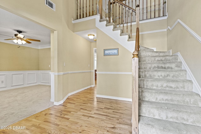 staircase featuring visible vents, a towering ceiling, a ceiling fan, wainscoting, and wood finished floors