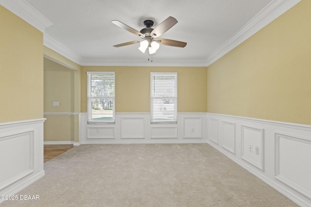 empty room with a ceiling fan, a wainscoted wall, light carpet, and crown molding