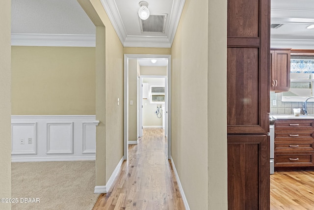 corridor featuring a wainscoted wall, crown molding, visible vents, a decorative wall, and light wood-style floors