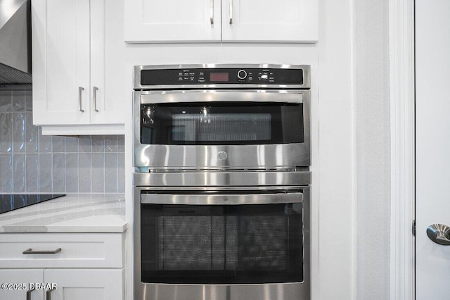 kitchen with tasteful backsplash, white cabinetry, light stone countertops, stainless steel double oven, and wall chimney exhaust hood