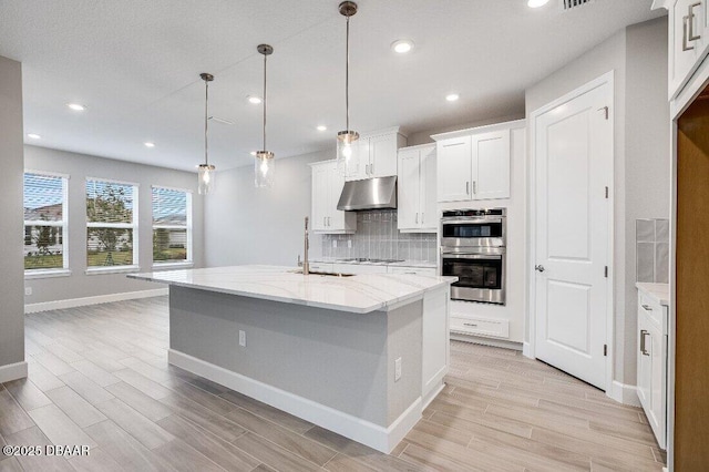 kitchen featuring white cabinetry, tasteful backsplash, an island with sink, decorative light fixtures, and stainless steel double oven