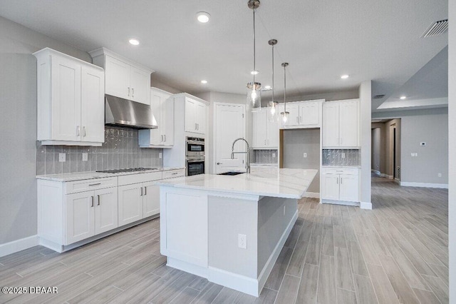 kitchen featuring double oven, decorative light fixtures, an island with sink, sink, and white cabinets