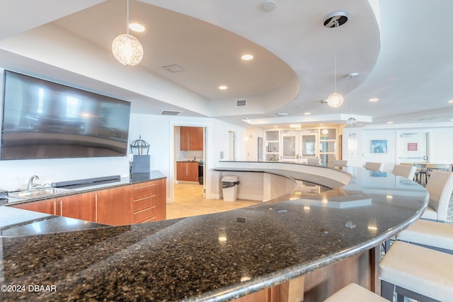 kitchen featuring a kitchen breakfast bar, dark stone countertops, a raised ceiling, sink, and decorative light fixtures