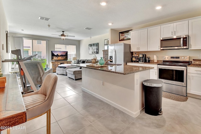 kitchen featuring appliances with stainless steel finishes, dark stone counters, a kitchen island with sink, ceiling fan, and white cabinetry