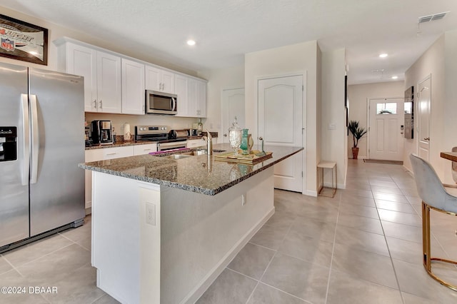 kitchen with stainless steel appliances, white cabinetry, a center island with sink, and dark stone countertops