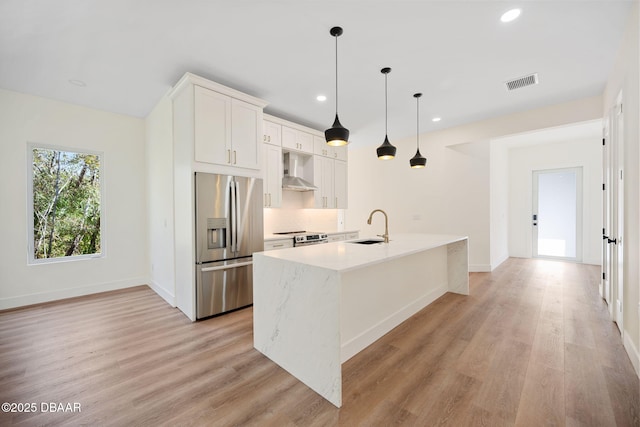 kitchen featuring wall chimney range hood, a kitchen island with sink, stainless steel appliances, white cabinets, and decorative light fixtures