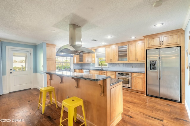 kitchen with appliances with stainless steel finishes, island range hood, a kitchen breakfast bar, light wood-style floors, and glass insert cabinets