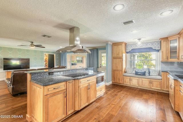 kitchen with black electric stovetop, visible vents, island exhaust hood, wallpapered walls, and dark stone counters