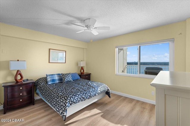 bedroom with ceiling fan, light hardwood / wood-style floors, a textured ceiling, and a water view