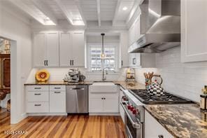 kitchen with stainless steel appliances, wall chimney range hood, beam ceiling, light hardwood / wood-style flooring, and white cabinets