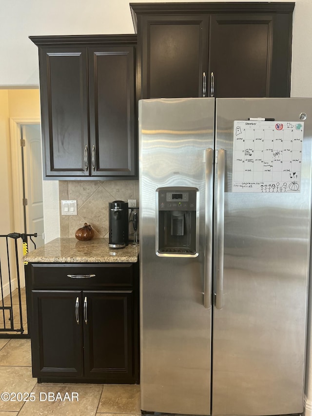 kitchen featuring light stone countertops, decorative backsplash, dark brown cabinetry, light tile patterned floors, and stainless steel fridge with ice dispenser