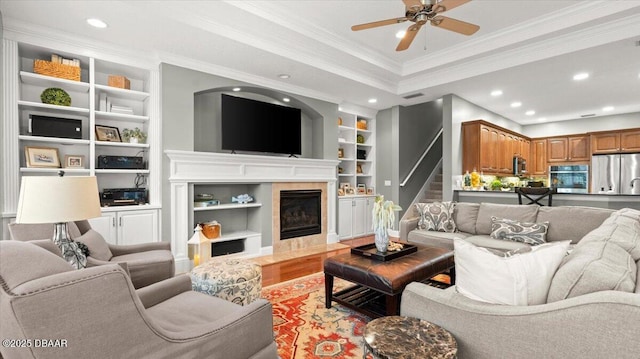 living room with built in shelves, crown molding, wood-type flooring, a tile fireplace, and ceiling fan