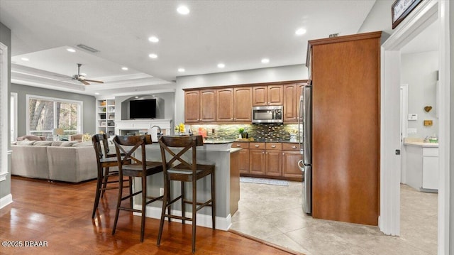 kitchen featuring appliances with stainless steel finishes, backsplash, a kitchen bar, a center island with sink, and a raised ceiling