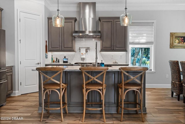 kitchen with a kitchen bar, wall chimney range hood, an island with sink, and light stone countertops