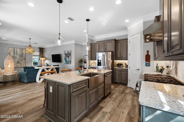 kitchen with stainless steel appliances, a kitchen island with sink, wall chimney range hood, dark brown cabinets, and decorative light fixtures