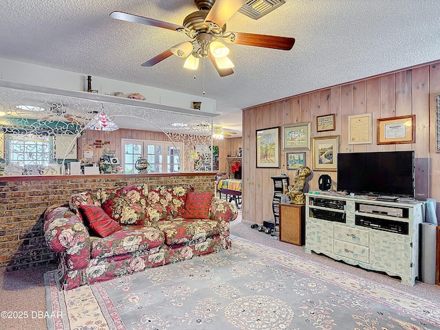 carpeted living area with visible vents, a textured ceiling, wooden walls, and a ceiling fan