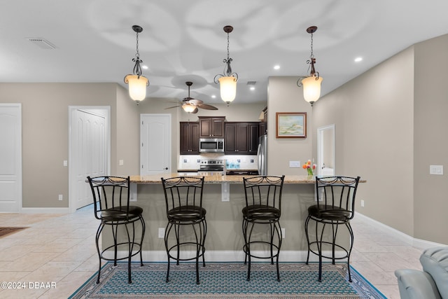 kitchen featuring dark brown cabinetry, ceiling fan, stainless steel appliances, light stone counters, and a breakfast bar area