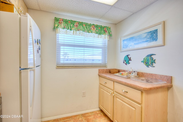 interior space with a paneled ceiling, light tile patterned flooring, light brown cabinets, and white fridge