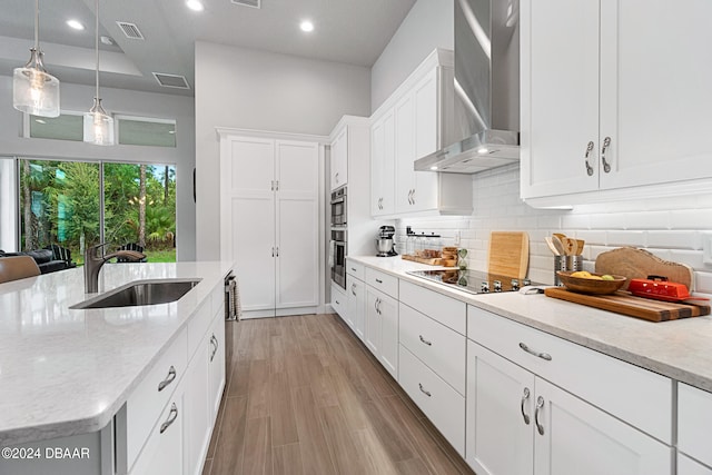 kitchen with black electric cooktop, hanging light fixtures, sink, wall chimney range hood, and white cabinetry