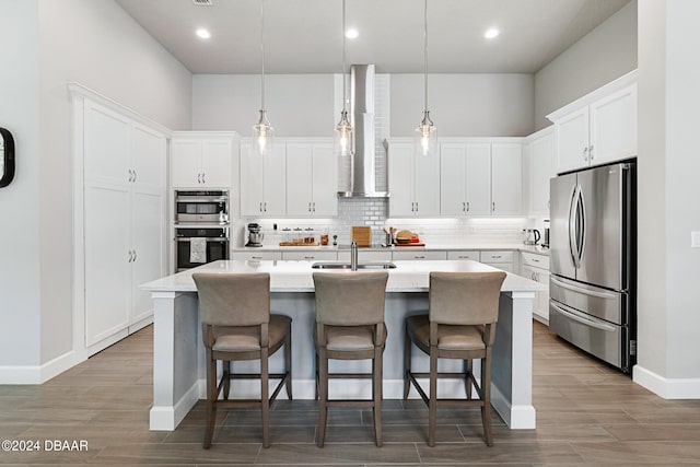 kitchen featuring white cabinetry, appliances with stainless steel finishes, pendant lighting, wall chimney exhaust hood, and a kitchen island with sink