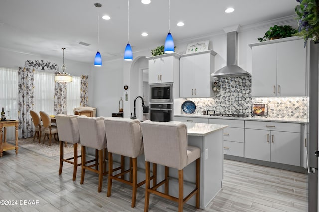 kitchen featuring white cabinets, appliances with stainless steel finishes, decorative light fixtures, a kitchen island with sink, and wall chimney range hood