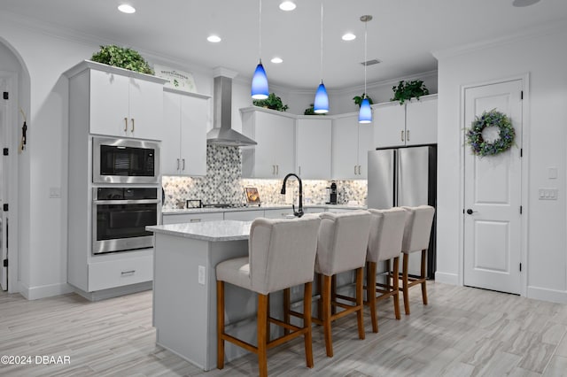 kitchen featuring an island with sink, wall chimney exhaust hood, appliances with stainless steel finishes, white cabinetry, and pendant lighting