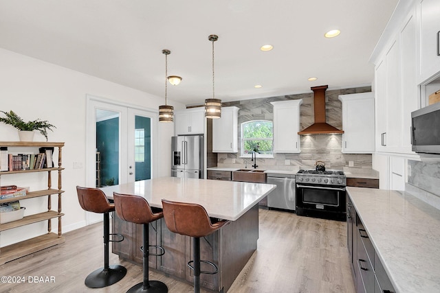 kitchen with white cabinets, wall chimney range hood, a kitchen island, appliances with stainless steel finishes, and decorative light fixtures