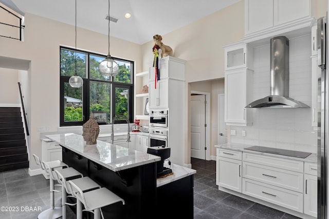 kitchen with light stone countertops, hanging light fixtures, a kitchen island with sink, white cabinets, and wall chimney range hood