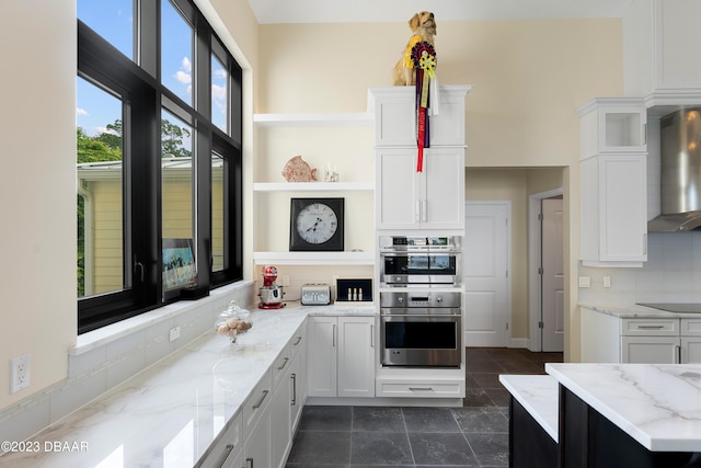 kitchen featuring white cabinetry, wall chimney range hood, stainless steel double oven, and light stone countertops