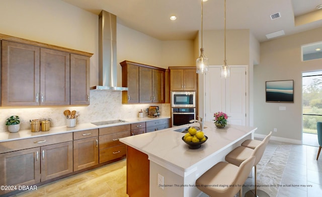 kitchen with wall chimney exhaust hood, hanging light fixtures, an island with sink, a breakfast bar, and appliances with stainless steel finishes