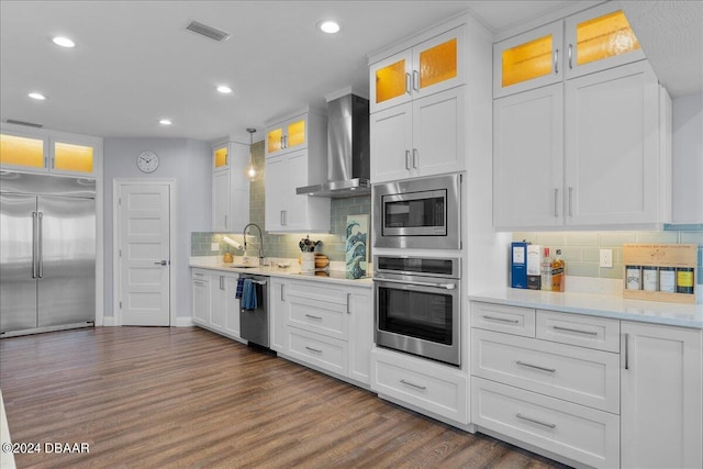 kitchen featuring wall chimney range hood, built in appliances, hanging light fixtures, white cabinets, and dark wood-type flooring