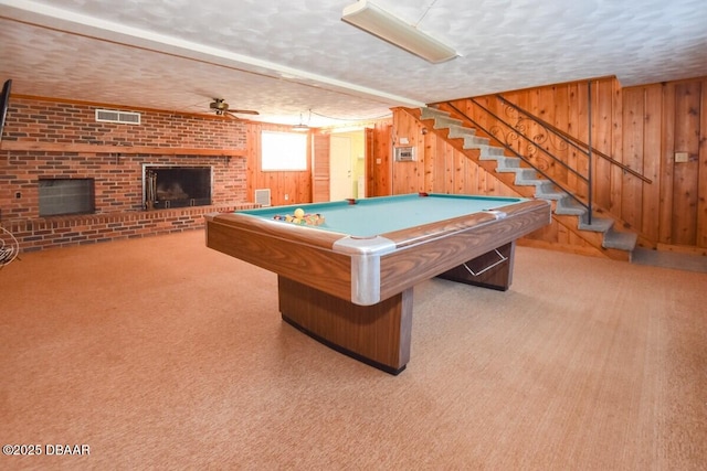 playroom with light colored carpet, a brick fireplace, a textured ceiling, wooden walls, and ceiling fan