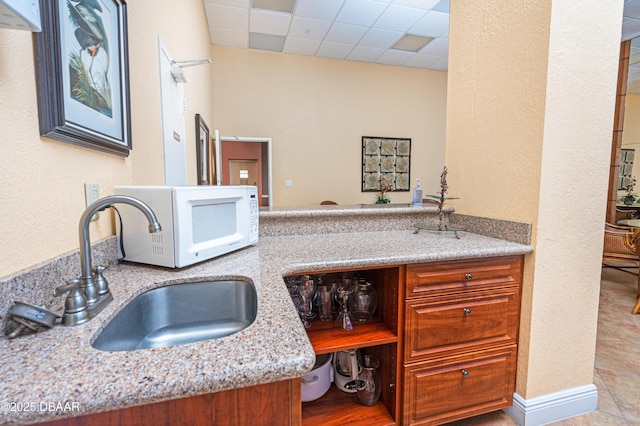 kitchen with white microwave, a drop ceiling, light stone counters, brown cabinets, and a sink