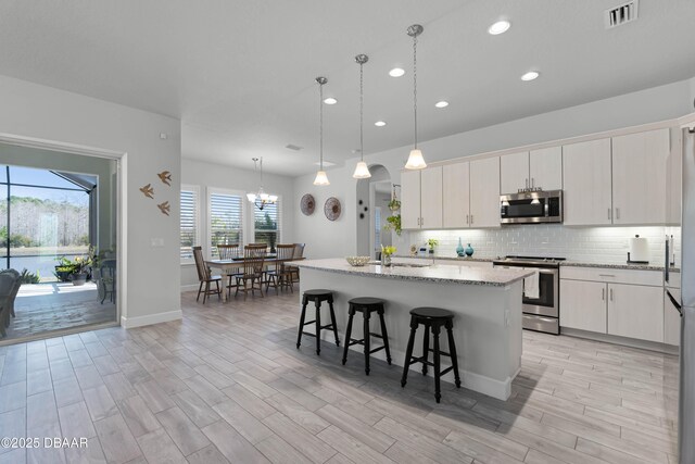 kitchen featuring white cabinetry, a center island with sink, stainless steel appliances, decorative backsplash, and a notable chandelier