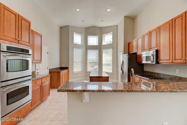 kitchen featuring kitchen peninsula, appliances with stainless steel finishes, dark stone countertops, light tile patterned floors, and a towering ceiling