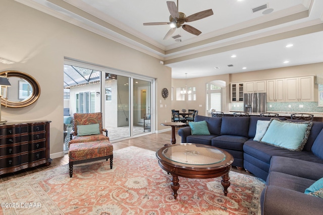 living room featuring ceiling fan with notable chandelier, ornamental molding, a raised ceiling, and light hardwood / wood-style flooring